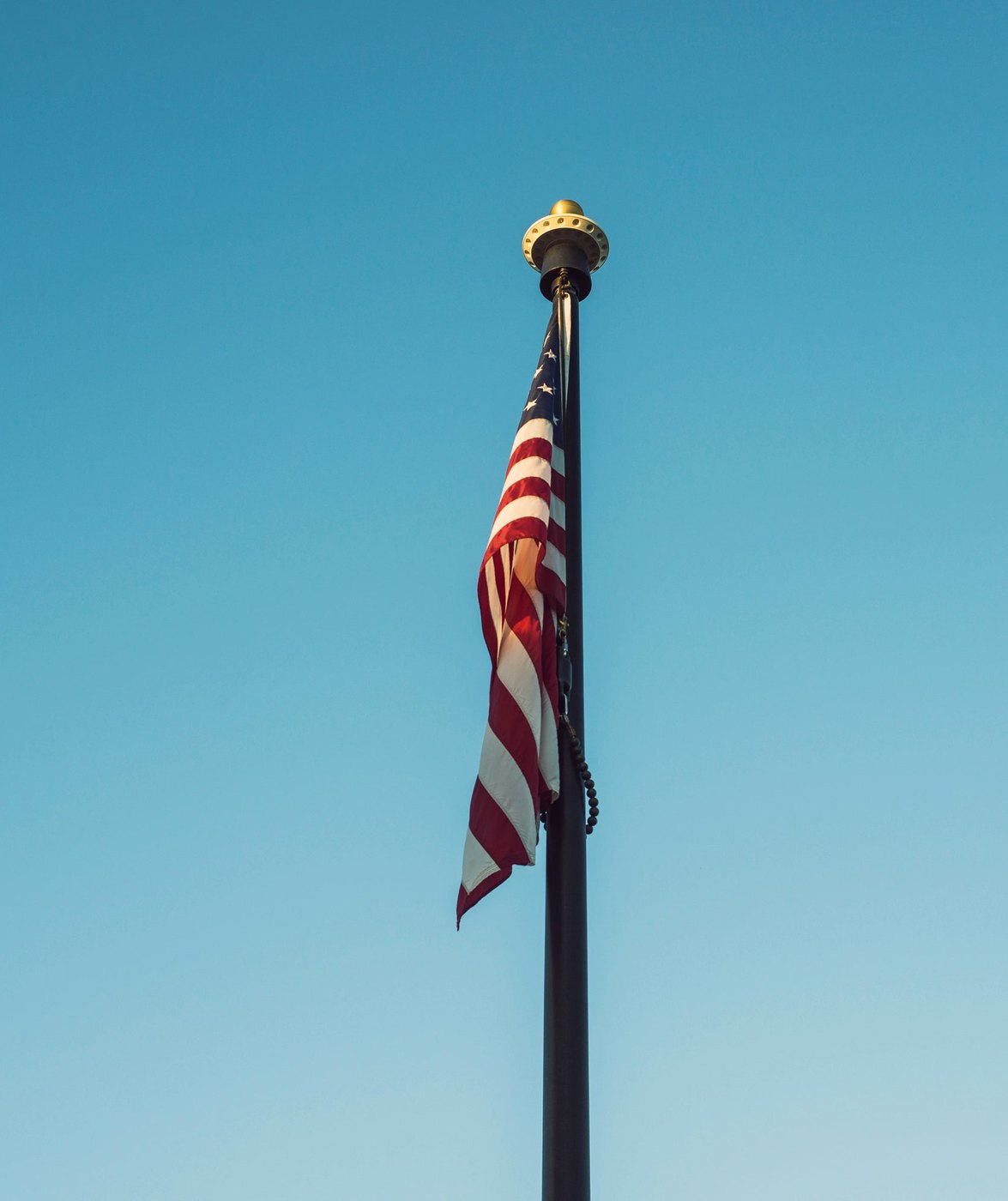An American flag flying from a tall dark flagpole with a gold ball on top, against a clear blue sky