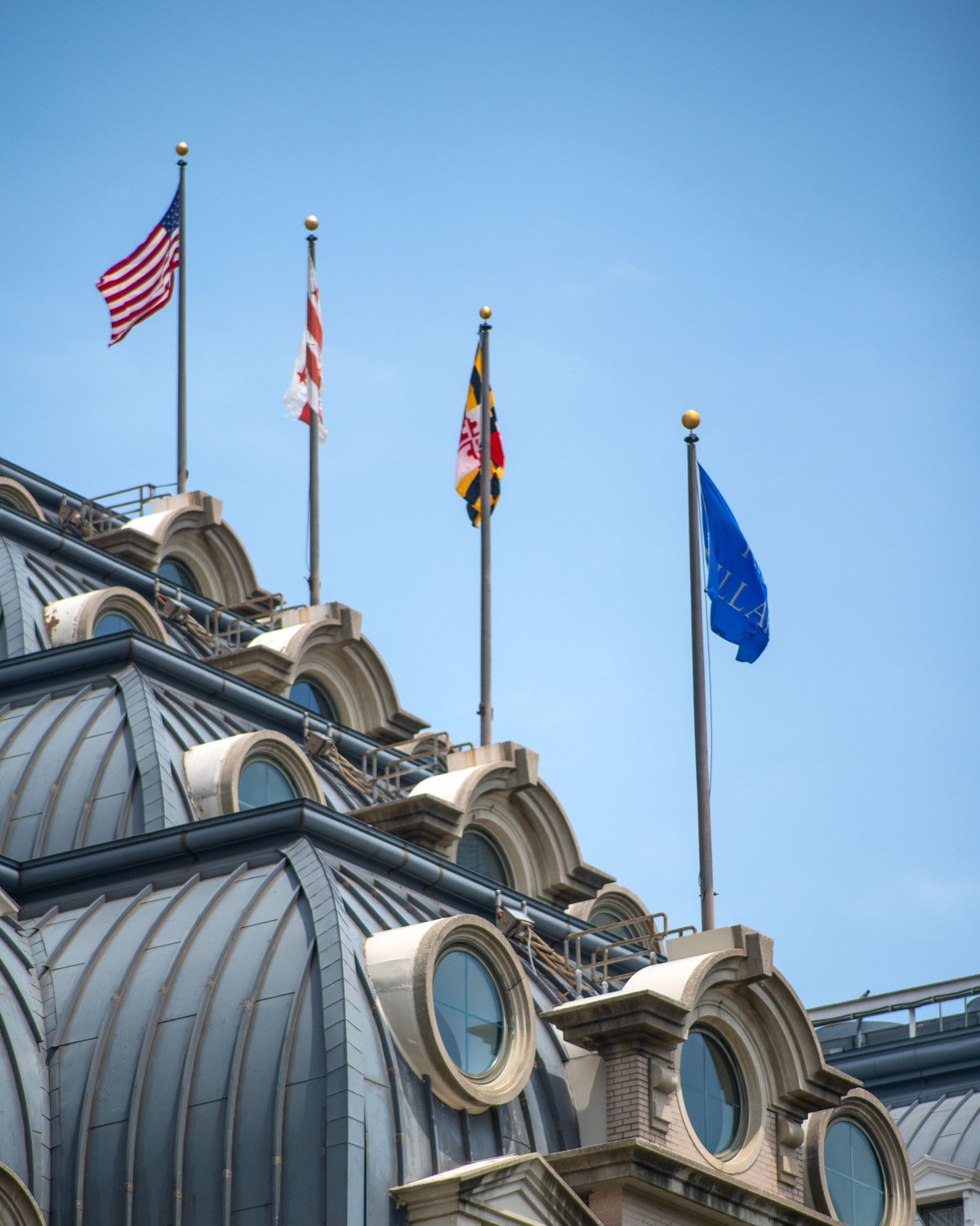 Four flags flying from a rooftop row of poles on a city building